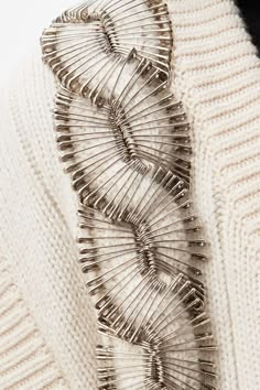 a closeup of a white sweater with safety pins sewn onto it in the shape of a flower. the sweater is soft and textured. the safety pins are shiny and metallic. the design is very intricate and detailed. this is a great example of how to add a unique touch to an everyday garment.