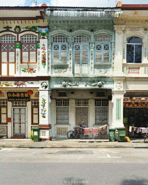 the image showcases a row of beautifully preserved shophouses in singapore.  the central shophouse is a striking light teal green, while the adjacent ones display a more vibrant, colorful style typical of peranakan architecture.  the buildings feature ornate details, including decorative carvings, intricate window frames, and colorful paintwork. a bicycle with laundry hanging on a line sits parked in front of the buildings, adding to the everyday scene captured. the street is quiet, bathed in the light of day.  the overall impression is one of vibrant history and cultural preservation within an urban environment.