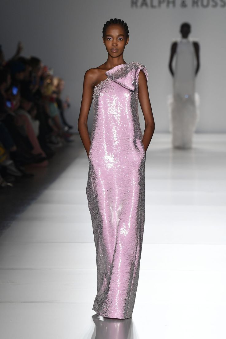 a model walks the runway at a fashion show wearing a oneshoulder pink and silver sequined gown. the dress is floorlength and has a loose, flowing fit. there are spectators in the background and a second model can be seen further down the runway. the backdrop includes the designers' brand name "ralph & russo".