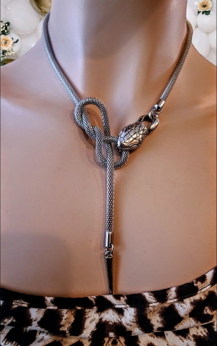 a closeup shot of a silver snake necklace on a mannequin, against a backdrop of blurry flowers. the necklace features a detailed snake head and a decorative spike at the end. the mannequin wears a leopard print top.