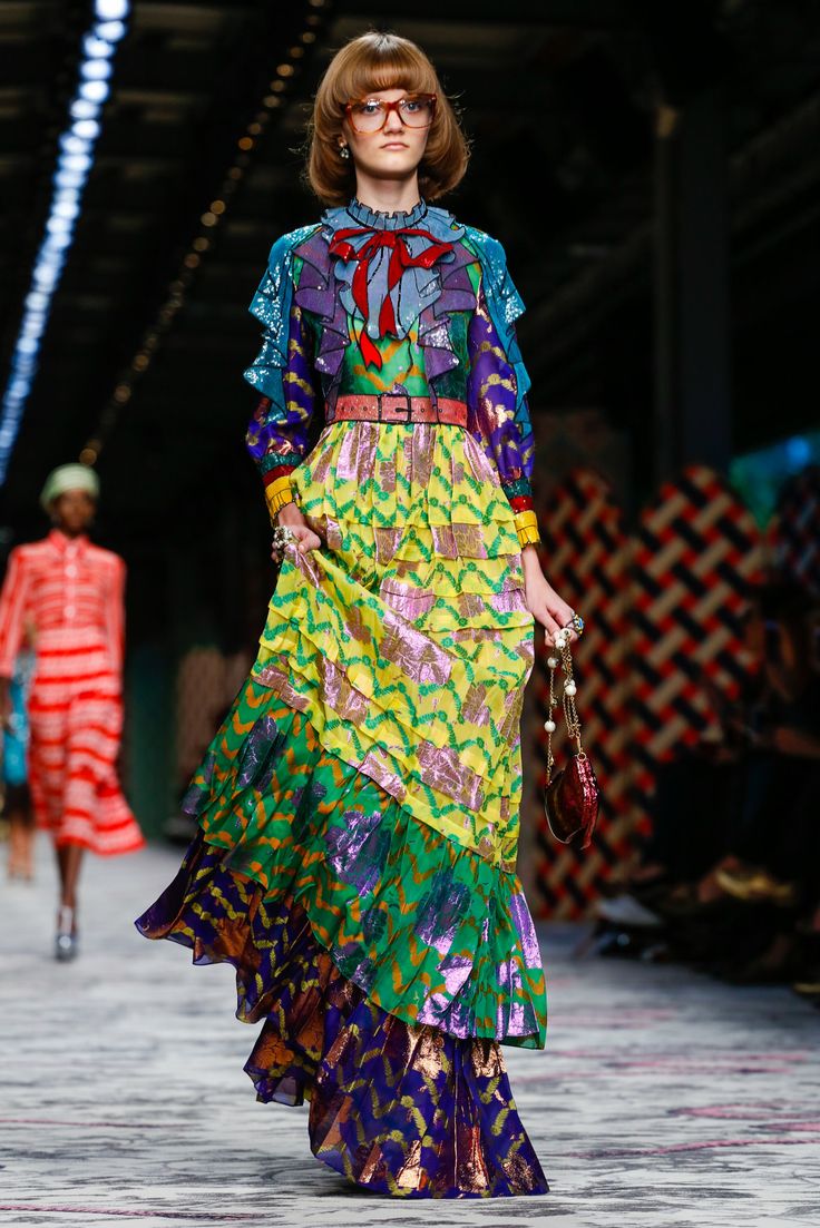 a model walks the runway at a gucci fashion show. she's wearing a long, multicolored maxi dress with a seventiesinspired pattern and ruffled details. the dress is predominantly yellow, green, and purple, and features a metallic sheen.  she also has on a red bow at the neckline, a belt, and is carrying a dark red handbag. her hair is a short, blunt bob, and she wears glasses.  another model wearing a red and white striped dress can be seen in the background, slightly out of focus. the runway is decorated with a geometric patterned floor.