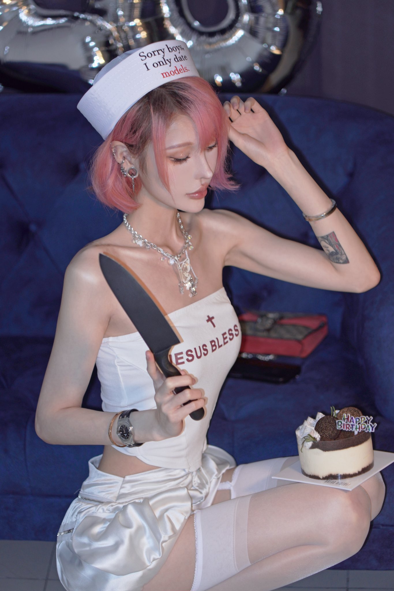 a young woman with pink hair, wearing a sailor hat that says "sorry boys i only date models", a white top that says "jesus bless", a white satin skirt, stockings, and a garter belt, sits on a couch holding a knife and looking at a small birthday cake.  the overall style is playful and slightly provocative.