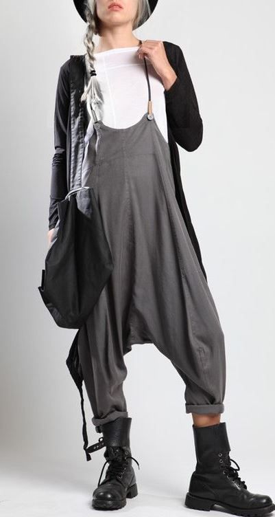 a woman is standing in front of a plain white background. she is wearing a white shirt under gray overalls, a long black cardigan, black boots, and a black hat. she has a braid in her hair. a black bag hangs off of her shoulder. the overalls have large pockets.