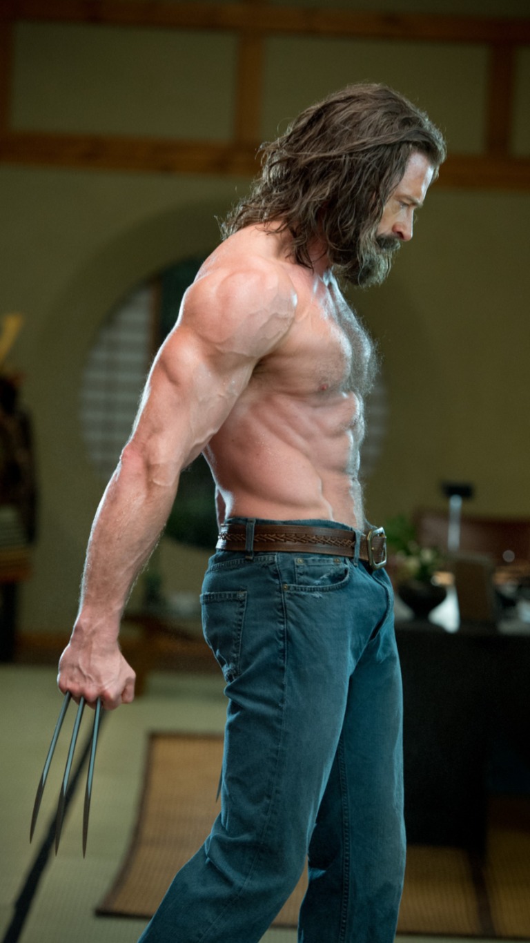 a side view of hugh jackman as wolverine, shirtless with long hair and a beard, wearing jeans and holding his claws. he is muscular and looking down.