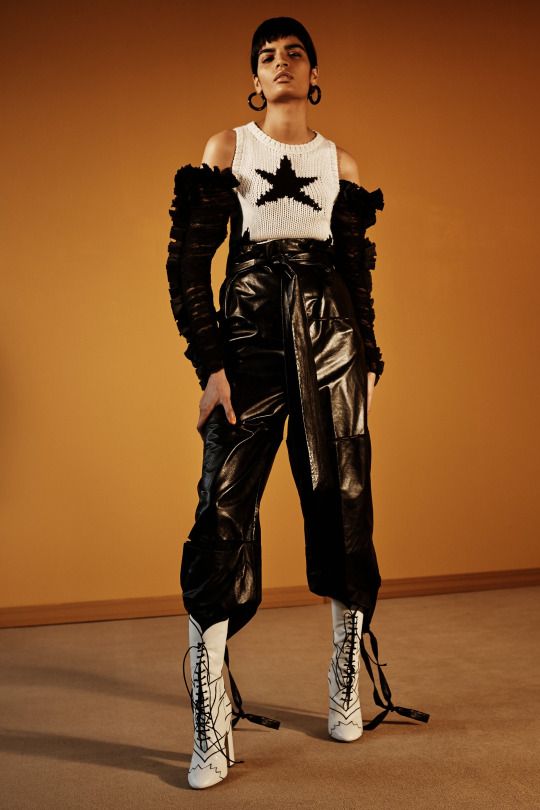 a woman poses in a studio wearing a white knit top with a black star, black leather pants, and white laceup boots. she is also wearing large hoop earrings. the background is a brown wall.