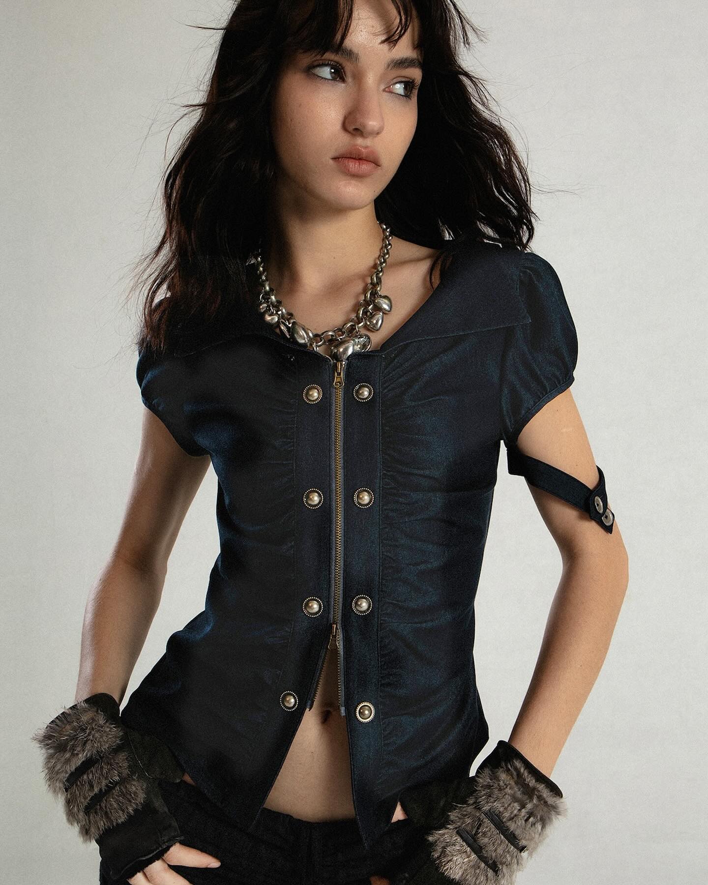 a young woman with long dark hair is modeling a dark blue shortsleeved shirt with a zipper and metallic buttons. she is wearing a chunky metallic necklace with heartshaped pendants and fur gloves. the shirt is slightly cropped, showing her midriff. she is posing against a light gray background.  the overall style is a mix of alternative, gothic and cyberpunk fashion influences.