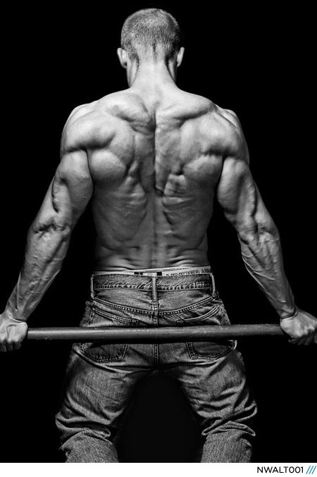black and white image of a muscular man's back as he holds a bar, likely in a gym or studio setting. the focus is on his defined back muscles and the lighting emphasizes his physique. ```