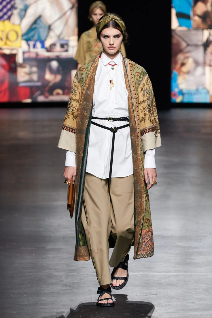 a fashion model walks the runway in a stylish outfit. she wears a long coat with a floral pattern over a white shirt and beige pants. she accessorizes with a wide belt, sandals, and a necklace. she's wearing a headband and has a small purse in her hand. the runway is a grey surface. there are graphic backgrounds behind her. the model is looking forward with a serious expression.