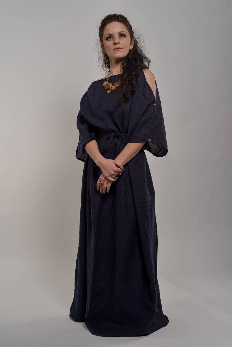 a fullbody studio shot of a woman wearing a dark blue linen maxi dress. the dress has offshoulder sleeves and a simple, loose fit. the woman has curly brown hair and is wearing a necklace and rings. she is standing against a plain gray background. the overall style is casual yet elegant.