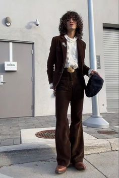 a young man in a brown corduroy suit stands on a sidewalk in front of a wall. he is wearing a white shirt, a gold belt, brown shoes, and a fedora. he is looking at the camera. the image is stylish and retro.