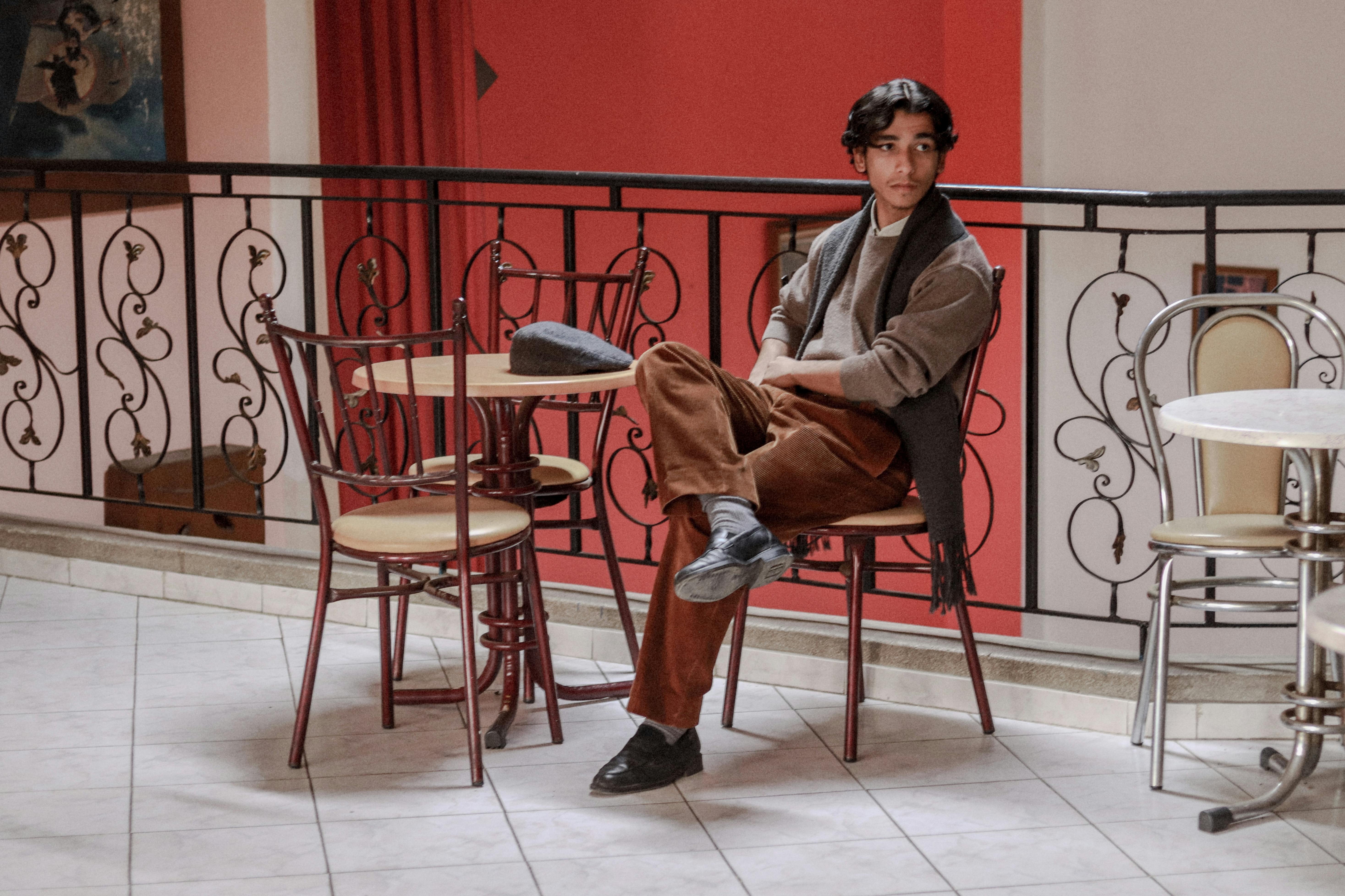 a young man with dark hair sits in a chair at a small table in a cafe. he is wearing brown corduroy pants, a light brown sweater, and a dark scarf. a hat rests on the table.  the cafe has a vintage feel, with metal chairs and tables, and a decorative balcony railing. the wall behind him is a deep red.  he appears relaxed and thoughtful.