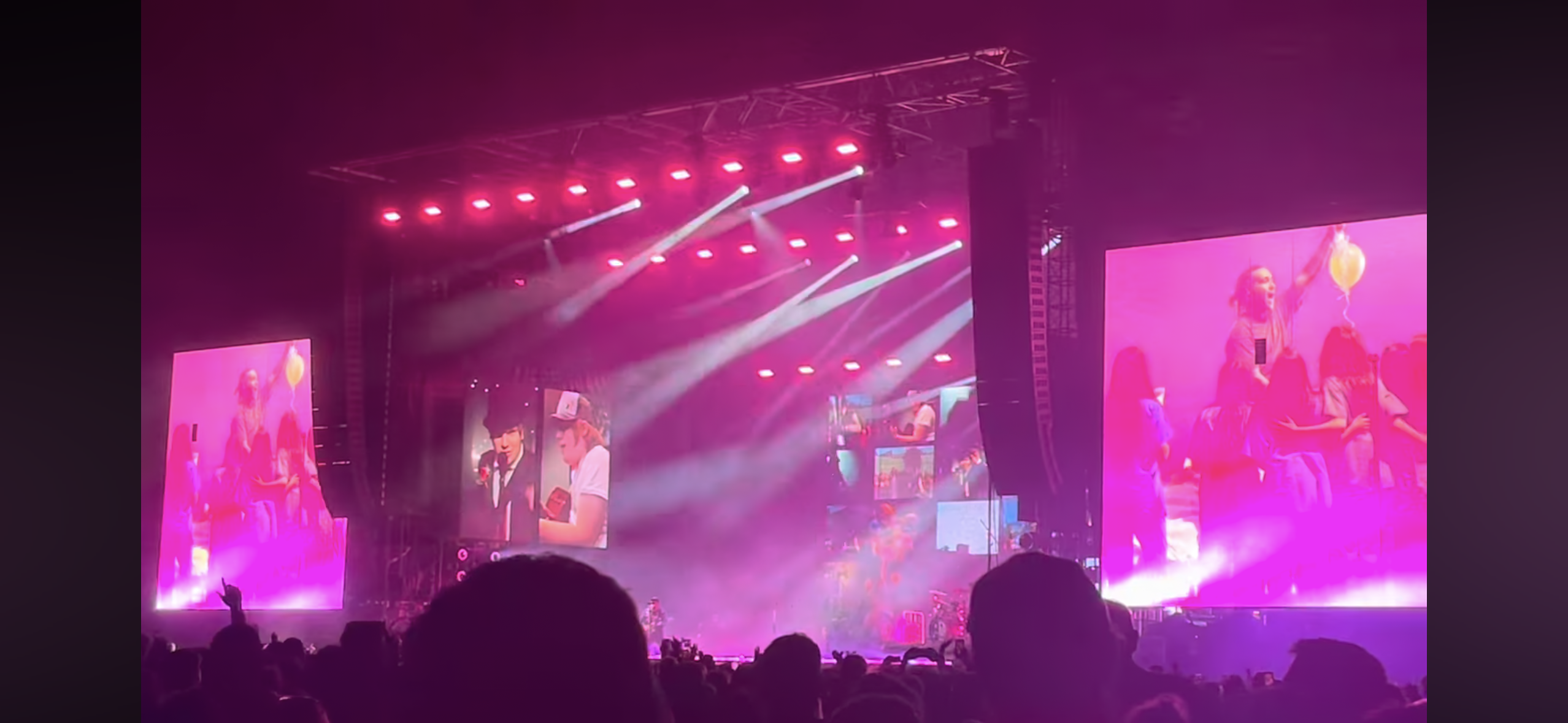 a concert stage with bright pink and white lights, the band is performing, and a large crowd is in the audience. there are two screens on each side showing the band performing. the stage is set up with a large video screen and sound equipment. the crowd is cheering and dancing along to the music.