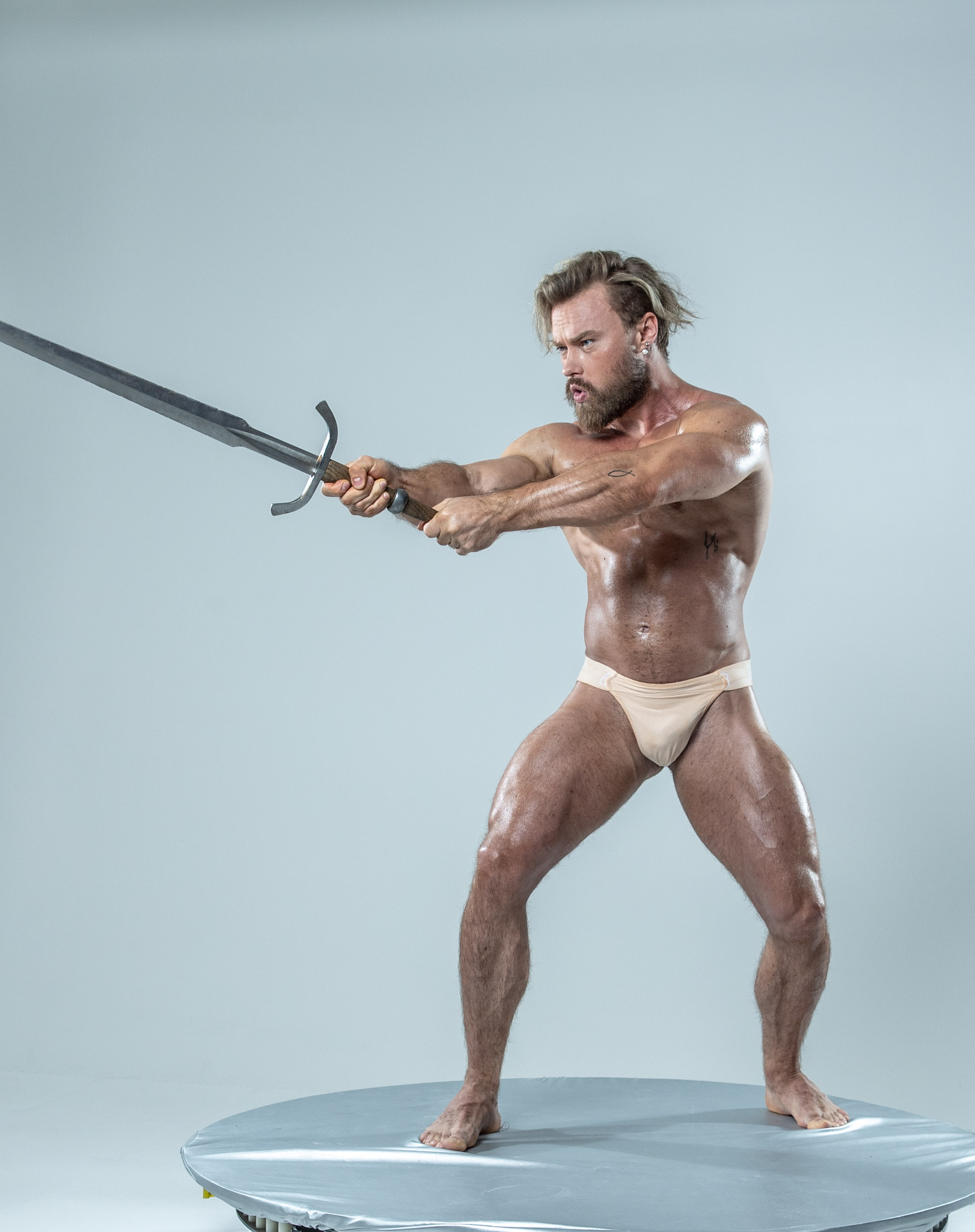 a muscular man with a beard and mustache is holding a sword with both hands in front of his body. he is in fighting stance with one leg in front of the other and his knees bent. the image is taken indoors in a studio with a white background. the man is lit with studio lighting.