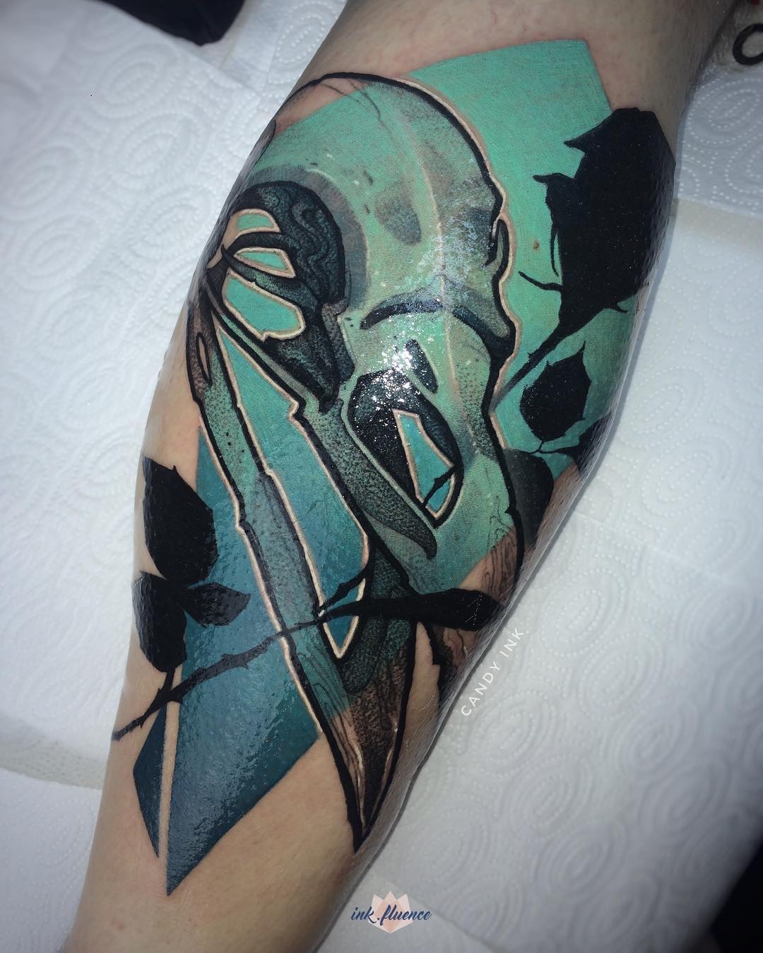 a tattoo of a bird skull, a rose, and abstract shapes in shades of teal, black and white on someone's forearm. the tattoo artist's signature "candy ink" is visible along with a watermark for "inkfluence." the photo appears to have been taken inside a tattoo parlor.