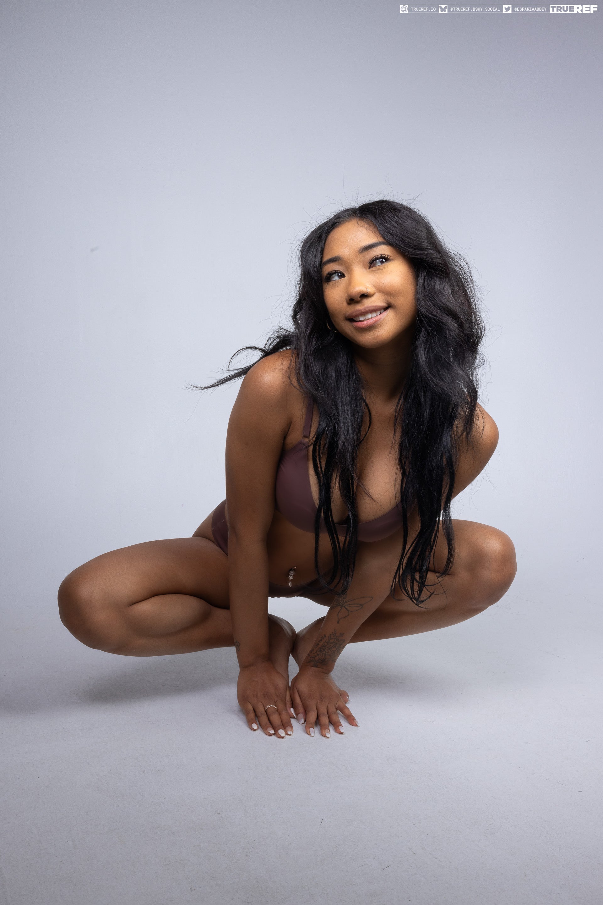 a woman is crouching in a studio shot, looking up and smiling. she has long black hair and is wearing swimwear. she has jewelry and a tattoo.