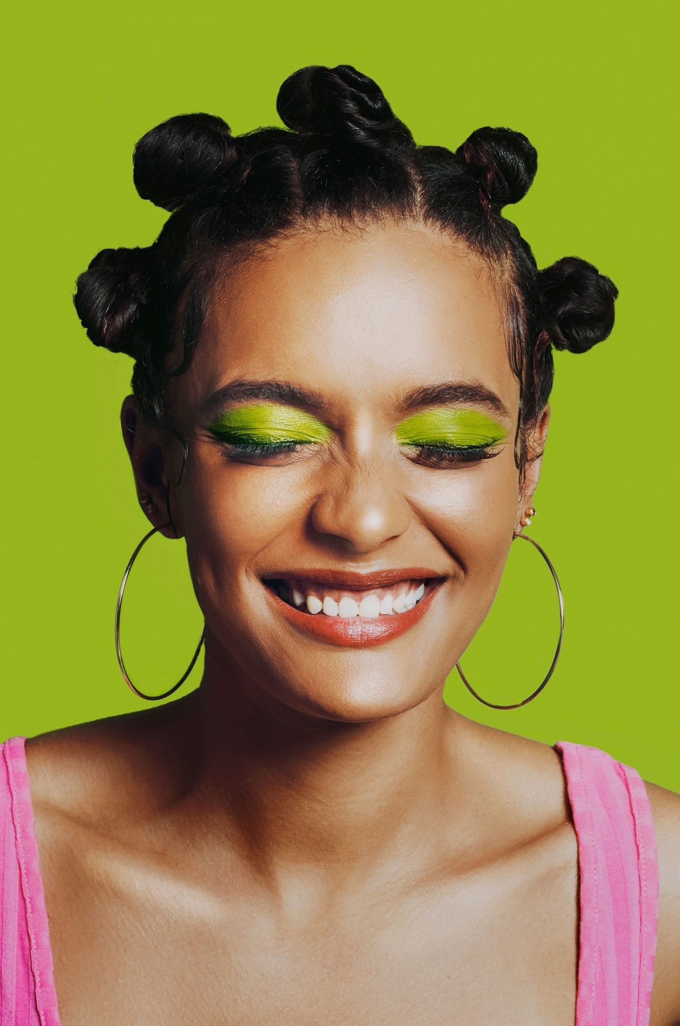 closeup portrait of a smiling black woman with vibrant green eyeshadow and her hair styled in bantu knots. she's wearing large hoop earrings and a pink camisole. the background is a bright lime green. the overall image is cheerful and stylish.
