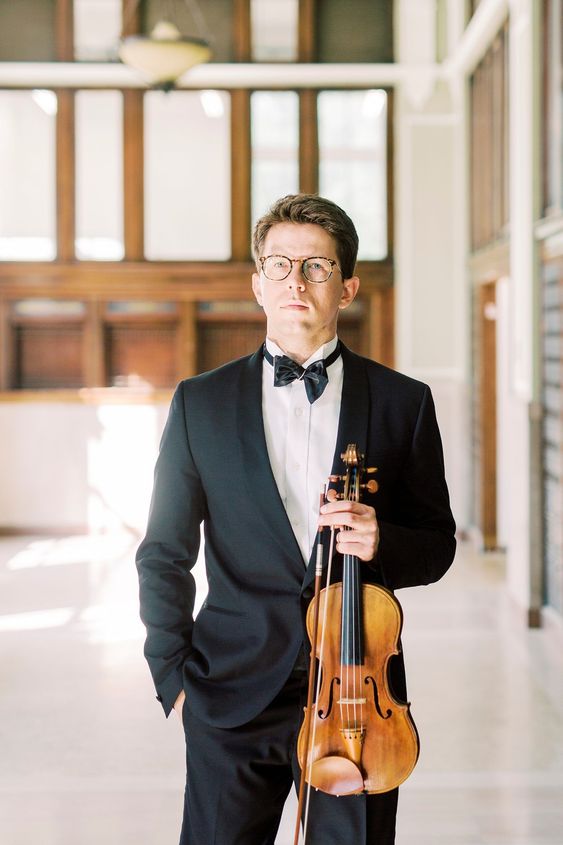 a man in a black suit and bowtie stands in a hallway, holding a violin and bow. he is wearing glasses and has a serious expression on his face. the hallway is bright and airy, with large windows letting in natural light.