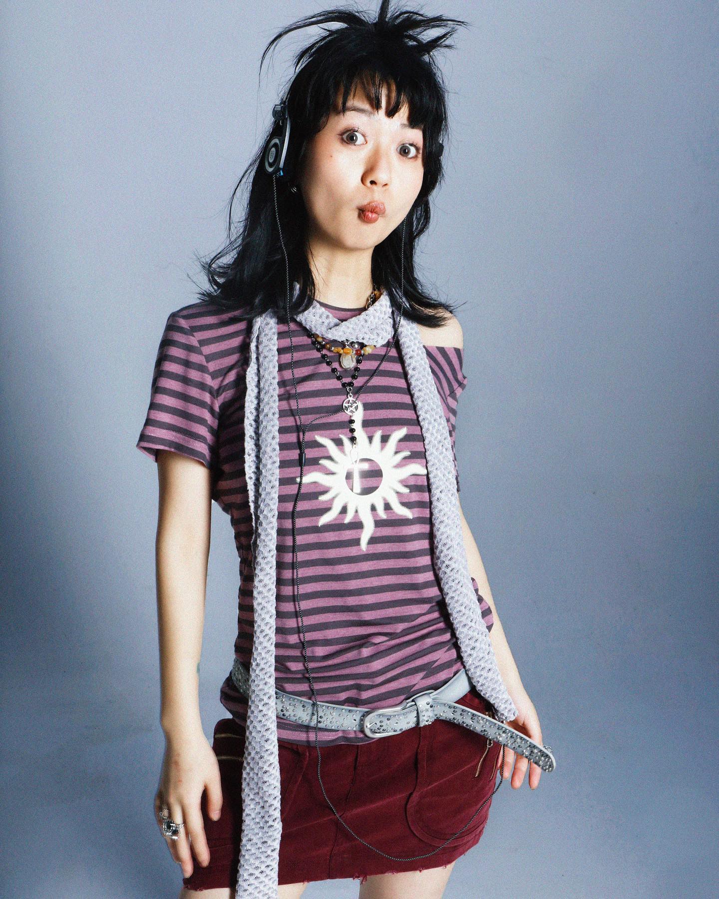 the image is a studio portrait of a young japanese woman, possibly a pop singer, styled in early 2000s punkpop fashion reminiscent of avril lavigne.  she's wearing headphones, a purple and grey striped tshirt with a sun design, a grey knitted scarf, a maroon corduroy mini skirt, and several necklaces. her dark hair is styled in a slightly messy, choppy bob. she's making a playful, slightly pouty facial expression. the overall aesthetic is casual yet trendy, reflecting the fashion sensibilities of early 2000s japanese pop culture and alternative styles.
