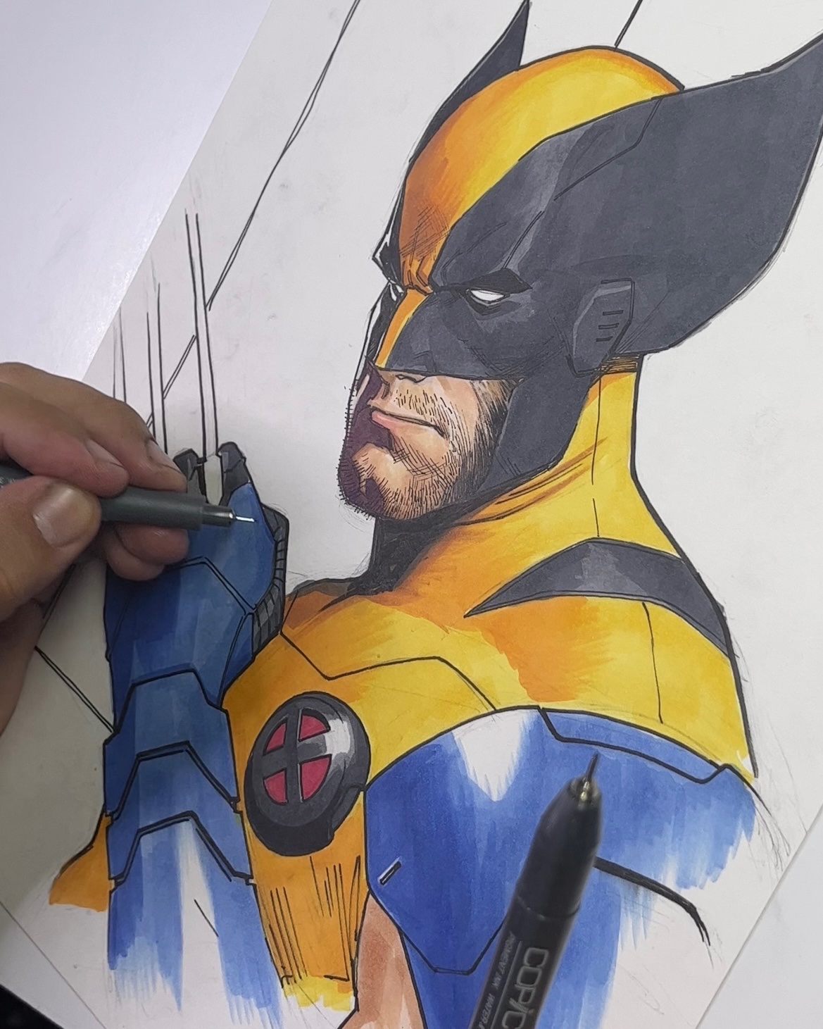 a hand holds a black pen and draws lines over a drawing of wolverine. the drawing is very detailed and features the superhero in a yellow and blue costume with black claws. wolverine's face is covered by a mask, and his eyes are partially visible. the drawing is on a white paper, and the artist is in the process of completing the artwork.  the drawing is likely a commissioned piece or a fan art project.