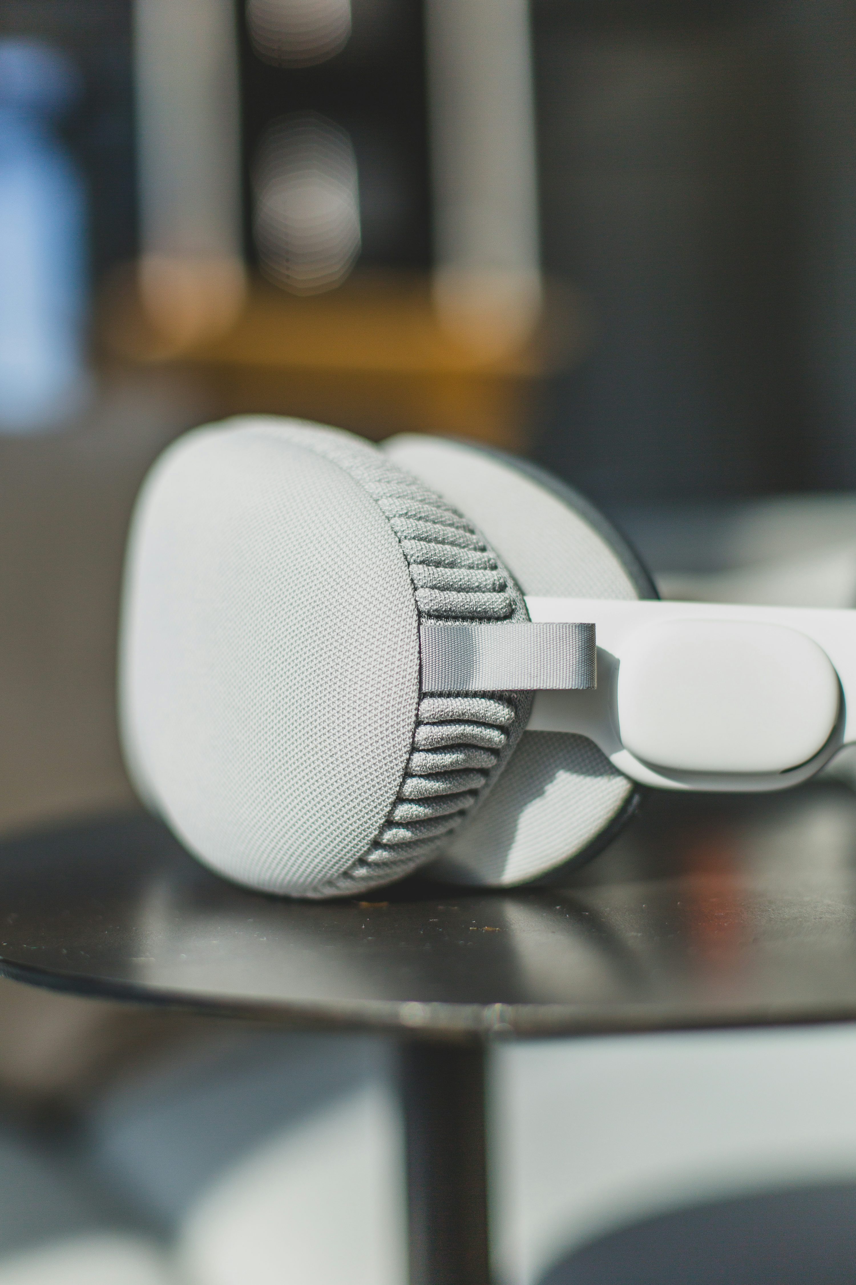 a closeup shot of a pair of grey and white headphones resting on a round, darkcolored table. the headphones have a fabric earpad and a visible headband. the background is slightly blurred, suggesting a shallow depth of field.