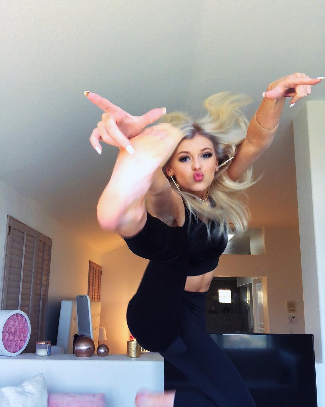 a woman with blonde hair is jumping in the air, wearing a black crop top and black leggings. she's pouting and pointing with both hands, and the photo appears to be a selfie taken indoors. in the background, there are various decorations, shutters, and a tv. she's wearing activewear.