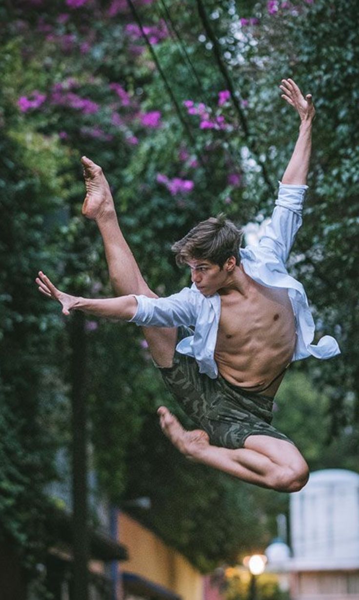 a male dancer, wearing a white shirt and camo shorts, leaps midair, with one leg extended to the side and his arms outstretched. he is in an urban park setting with trees, leaves, and flowers behind him. the dancer appears to be in midperformance.
