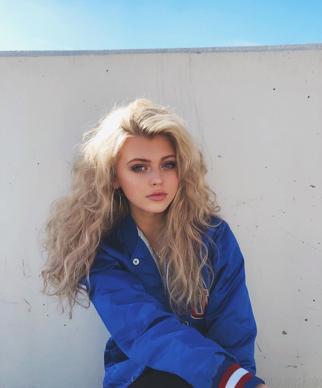 a young woman with long, wavy blonde hair stands in front of a white wall. she is wearing a blue jacket with red and white stripes on the sleeves. she has hoop earrings and is wearing naturallooking makeup. the sky is blue in the background.