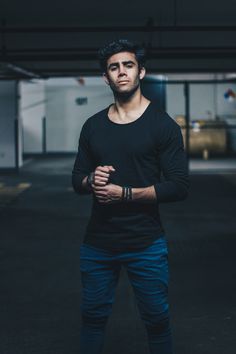 a young man with short dark hair and a serious expression stands in a dimly lit parking garage. he is wearing a black longsleeved shirt and blue jeans. the man is looking directly at the camera, his hands clasped in front of him. the background of the image is blurred, emphasizing the man's presence in the foreground.