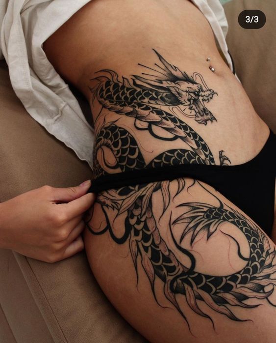 a woman is lying on a couch, revealing a large black dragon tattoo on her hip and thigh. she is wearing black underwear and a white top. a hand is pulling down the waistband of her underwear. her stomach is also visible, with a piercing near her belly button.