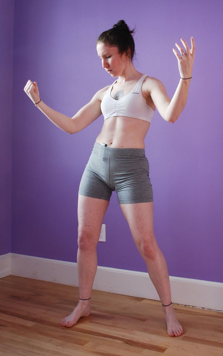 a woman in a white sports bra and grey shorts is standing barefoot on a wood floor, with her arms raised above her head and her palms facing up. she is looking down with a smile on her face. the wall behind her is purple. she is standing with her feet slightly apart and her knees are slightly bent. her hair is tied back in a ponytail. the woman has a happy and confident expression on her face.