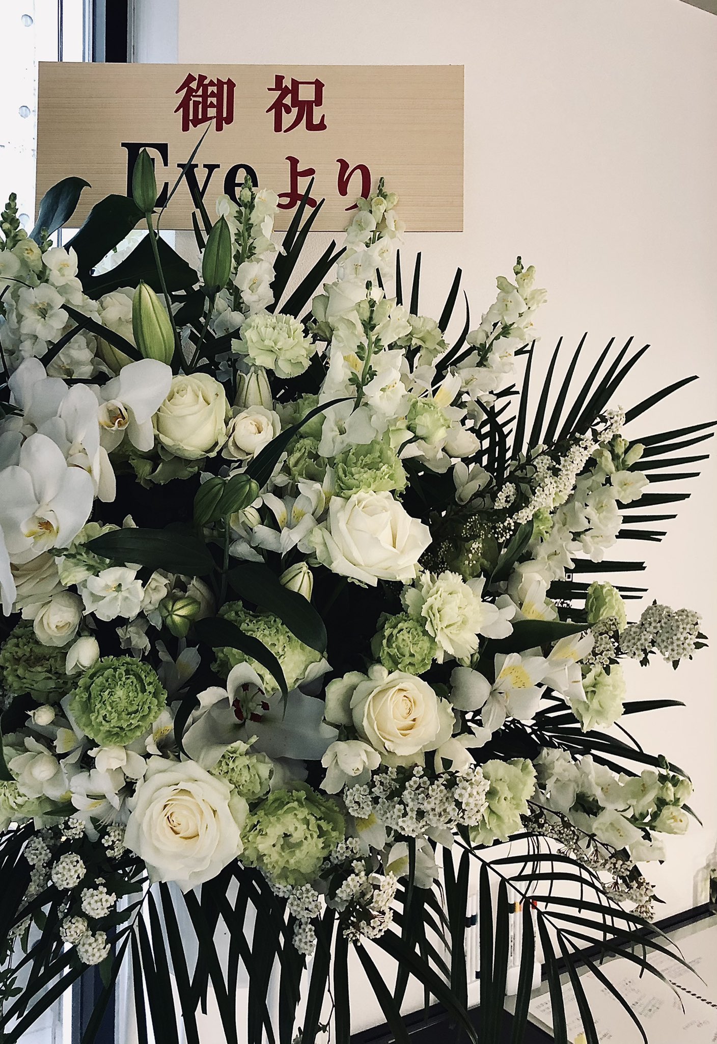 a large floral arrangement with white lilies, roses, and other green and white flowers. there is a wooden sign partially obscuring the arrangement that reads "eve" in english and has japanese characters above it.  the flowers appear to be for the opening of a business.