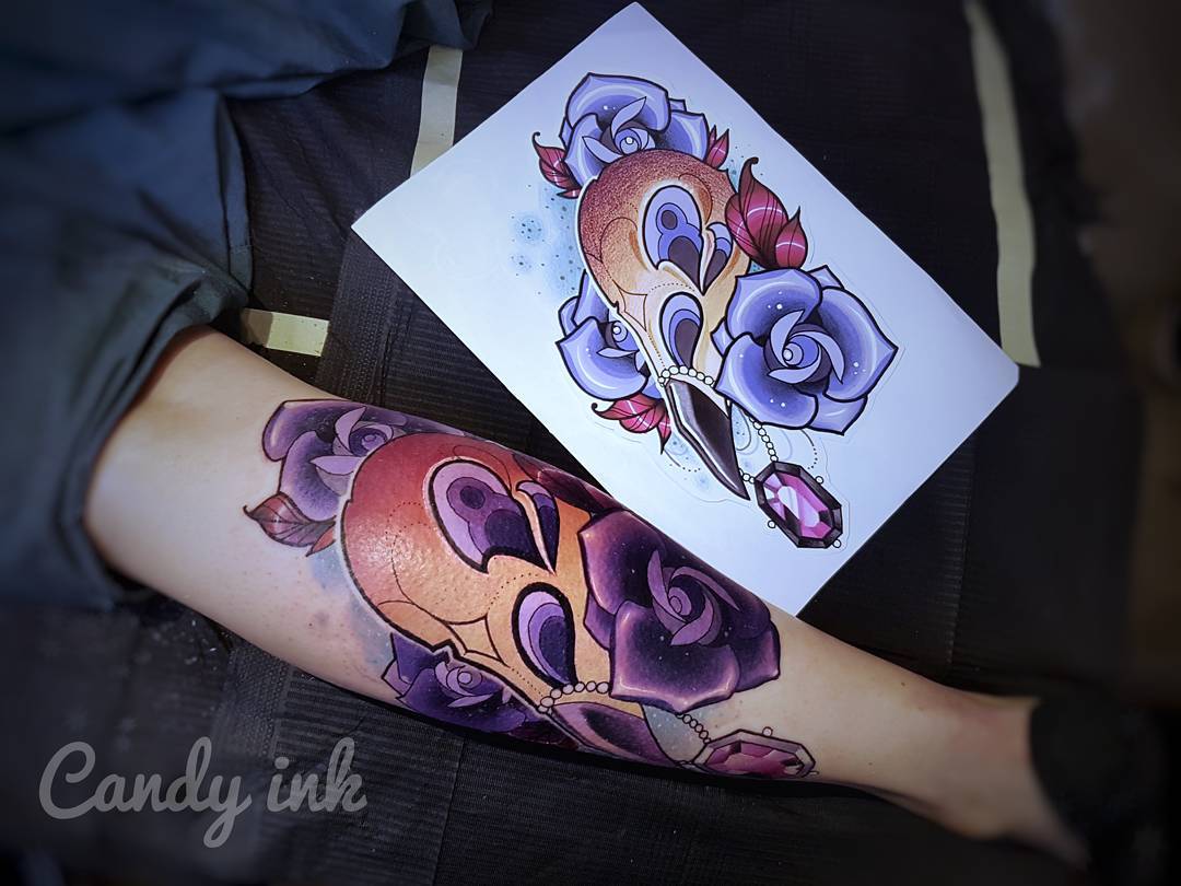 a person has gotten a neotraditional style tattoo of a swan skull with roses and a crystal on their inner forearm. there's a printed sketch of the tattoo design above the tattooed arm.