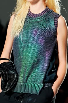 a woman in a sleeveless knit sweater with ombre green, blue and purple hues. she is wearing black pants and a black handbag.  her blonde hair is pulled back from her face and she is looking at the camera.