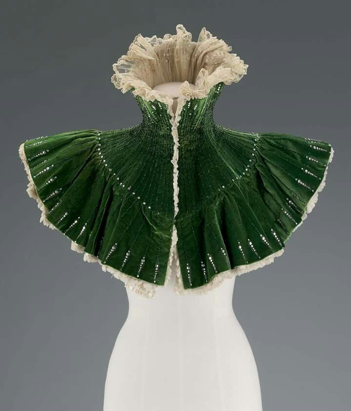 a green velvet cape with lace trim is displayed on a white mannequin. the cape has a ruffled collar and is decorated with small, shiny embellishments. the cape is open in the front.