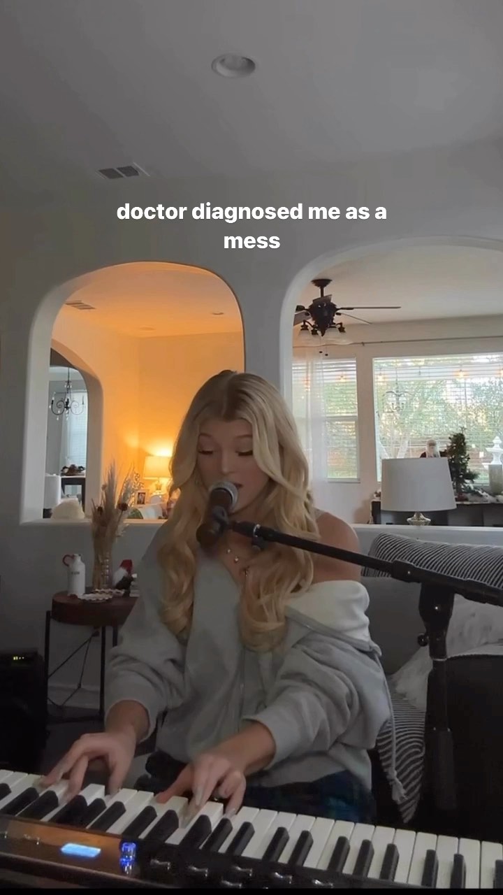 a young woman with blonde hair is sitting at a keyboard and singing into a microphone. she's wearing a casual grey sweater and appears to be in a home environment. the background shows part of a living room with arches. the text overlay on the image reads, "doctor diagnosed me as a mess".