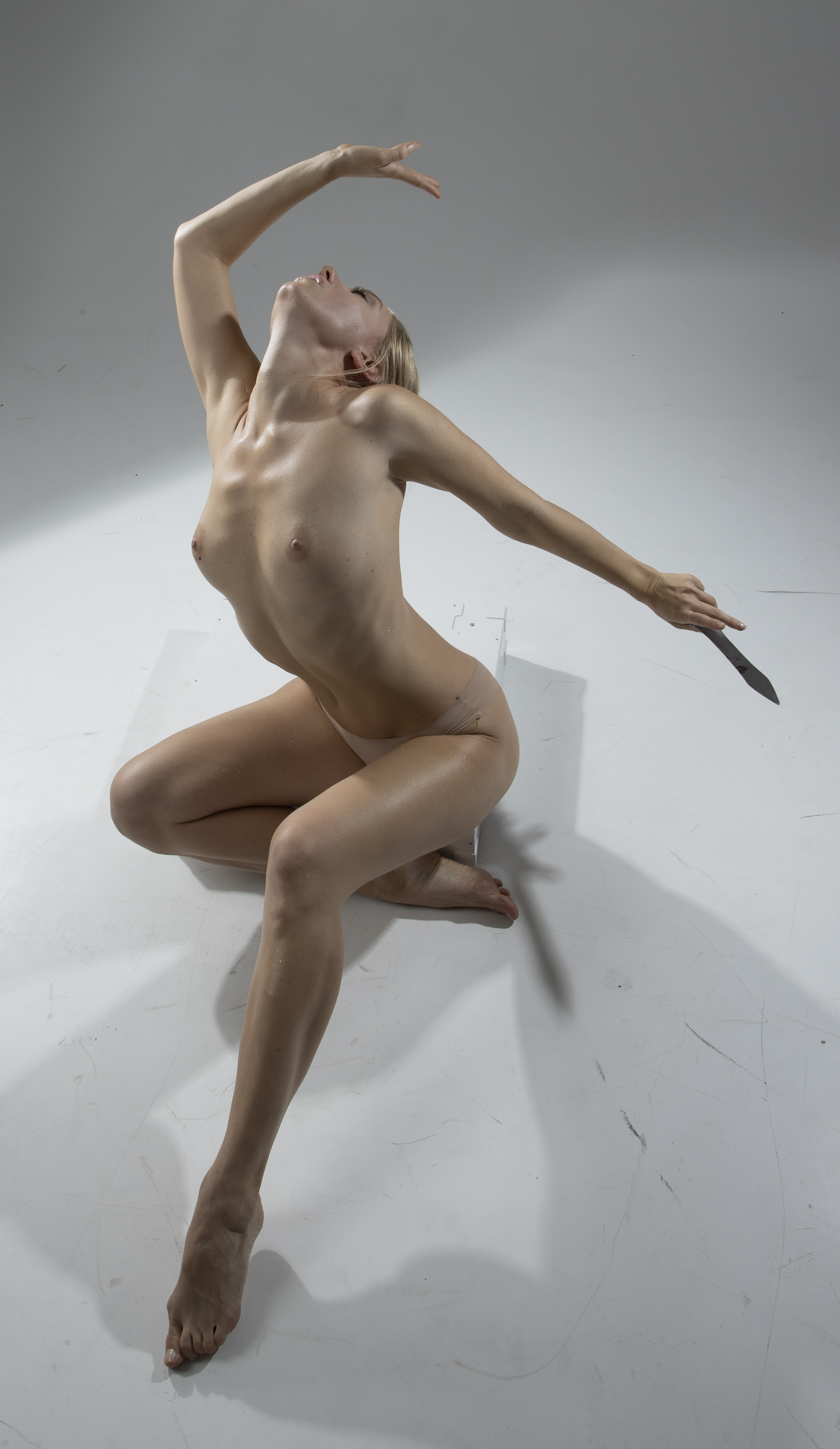 a fullbody studio shot of a naked woman with blonde hair, in an artistic pose, holding a knife, on a white background. the woman is looking upwards, with one arm raised and one leg bent, while she kneels on the other leg. the scene is illuminated with studio lighting, casting a shadow on the white background. the subject's pose highlights the female form in an anatomical manner.
