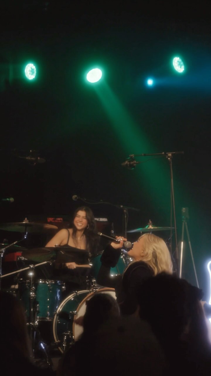 a photograph captures a live musical performance. a female drummer with long, dark hair smiles as she sits behind a blue drum set. beside her stands a blonde female singer, holding a microphone and singing. stage lighting shines down on the performers. the audience is visible in the foreground.