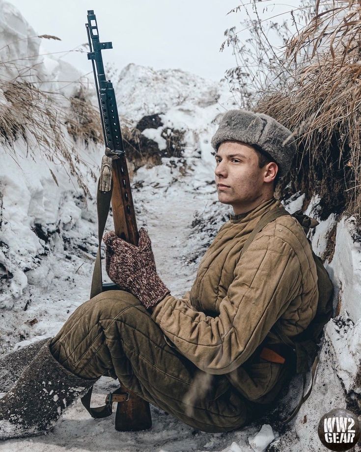 a soldier wearing a padded winter uniform and a fur hat sits in a snowy trench, holding a rifle, staring off into the distance. he's likely engaged in a reenactment of the battles of the eastern front during world war ii.
