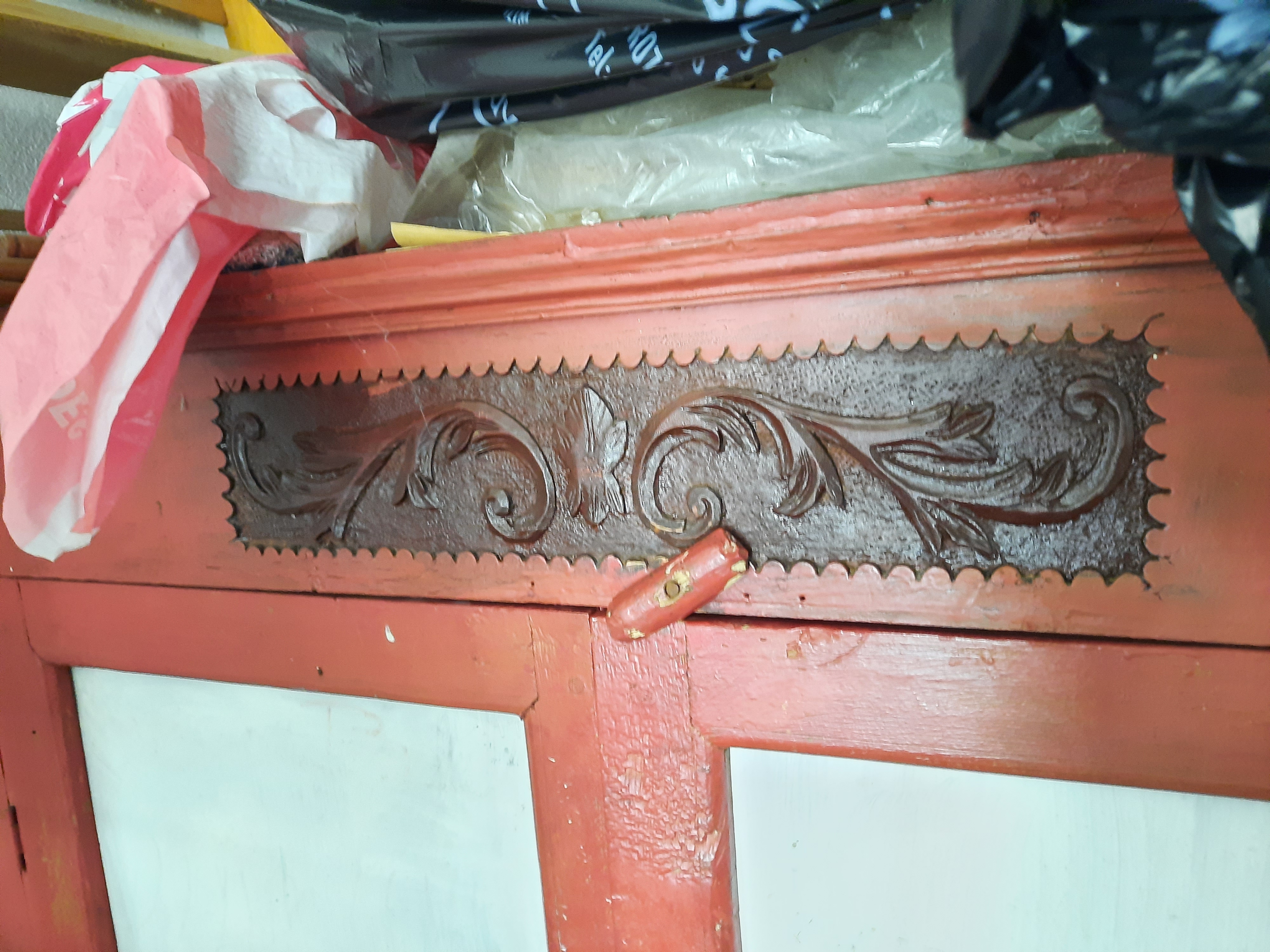 closeup shot of a wooden cabinet with a decorative carved wood panel above two whitepainted doors. the carved panel features a dark brown floral design against a red painted background. a red painted wooden latch secures the doors. a red and white plastic bag sits on top of the cabinet to the left, while other items covered in plastic bags are visible on top.