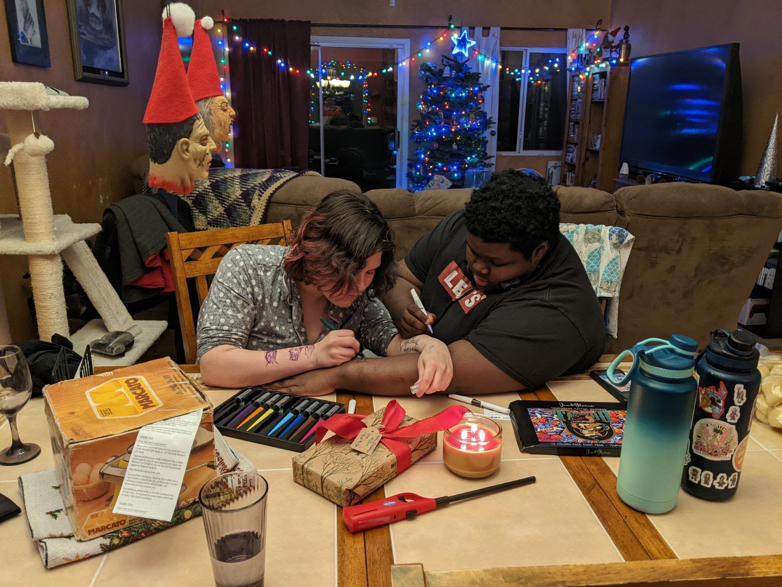a woman with dark hair and a black shirt is drawing a tattoo on a man's arm. the man is wearing a black shirt and is sitting at a table with his arm outstretched. the table is covered in tattoo supplies, including markers, stencils, and a lighter. there is a christmas tree in the background, and the room is decorated for the holidays.