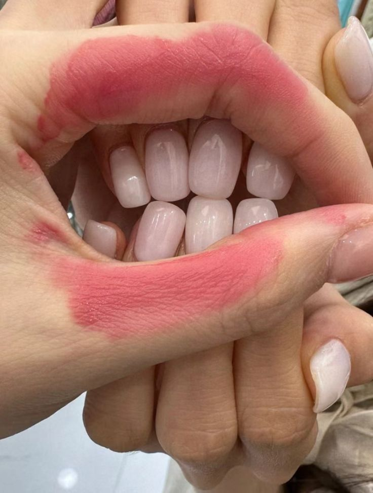 the image shows a closeup of a woman's smile, highlighting her bright white teeth. her lips are adorned with a rosy pink lipstick, and her hands, with manicured white nails, are framing her mouth. the bright, artificial lighting emphasizes the whiteness of her teeth and the vibrancy of the lipstick.