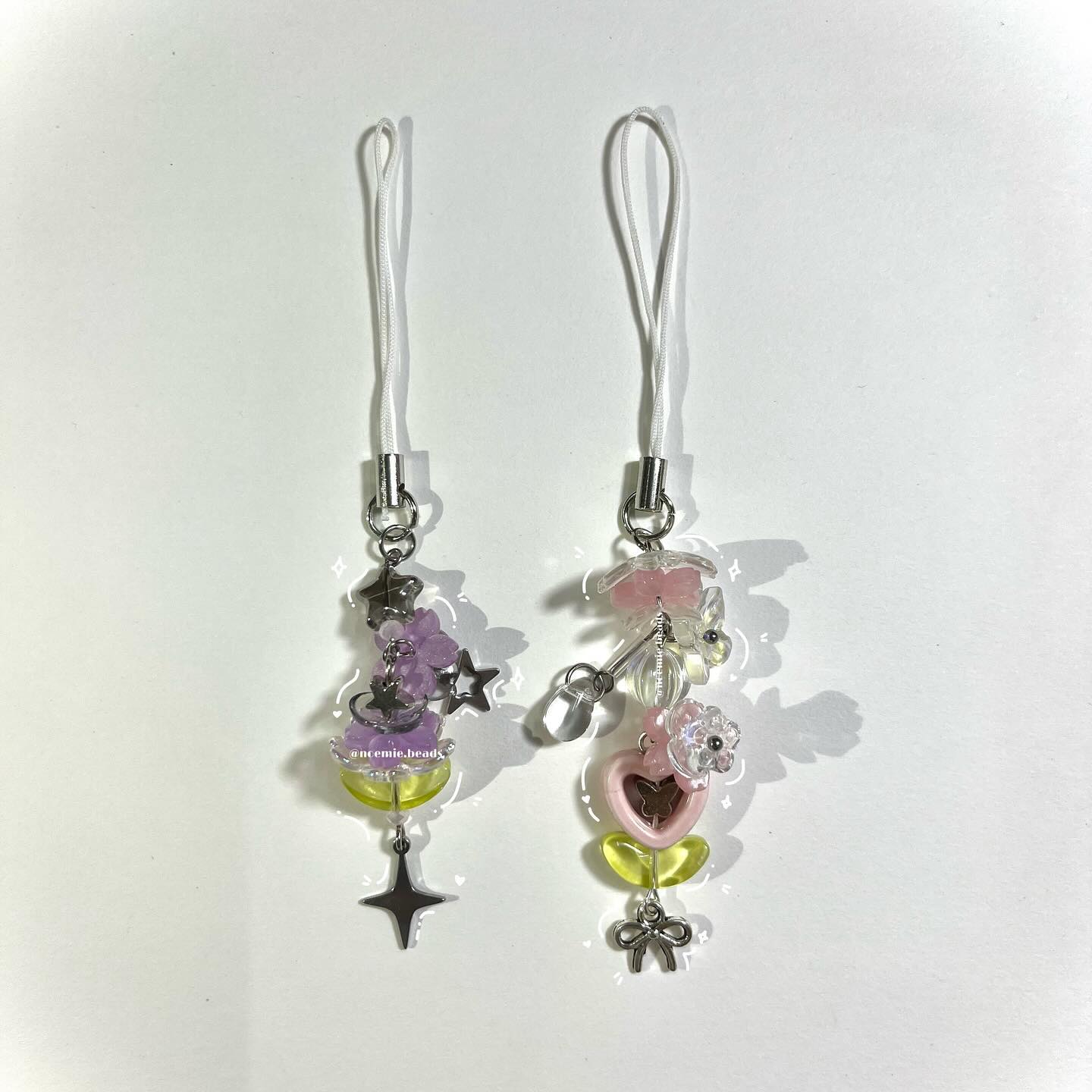 two beaded phone charms laying side by side on a white surface. the left charm has lavender and light green beads along with silver star charms and a silver cross charm at the bottom. the right charm has pink, clear, and light green beads, a heart shaped bead, flower beads, and a bow charm at the bottom. both charms have a white lanyard attached to silver hardware at the top.