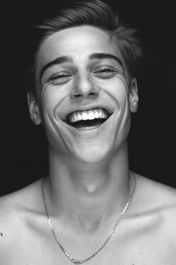 a black and white portrait of a young man smiling with a white necklace on.  the man has a bright smile and his teeth are very white.  he is looking directly at the camera and is dressed in casual clothing.  the image is welllit and the man is the only subject in the image.