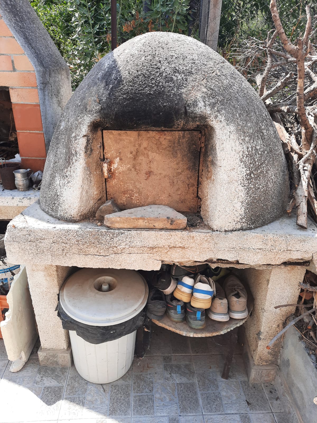 the image shows a stone oven with a metal door in an outdoor kitchen. there is a trash can and a shelf with shoes underneath the oven. a chimney is visible on the left side of the oven. twigs are piled on the right side of the oven. a bucket is on the left side on top.