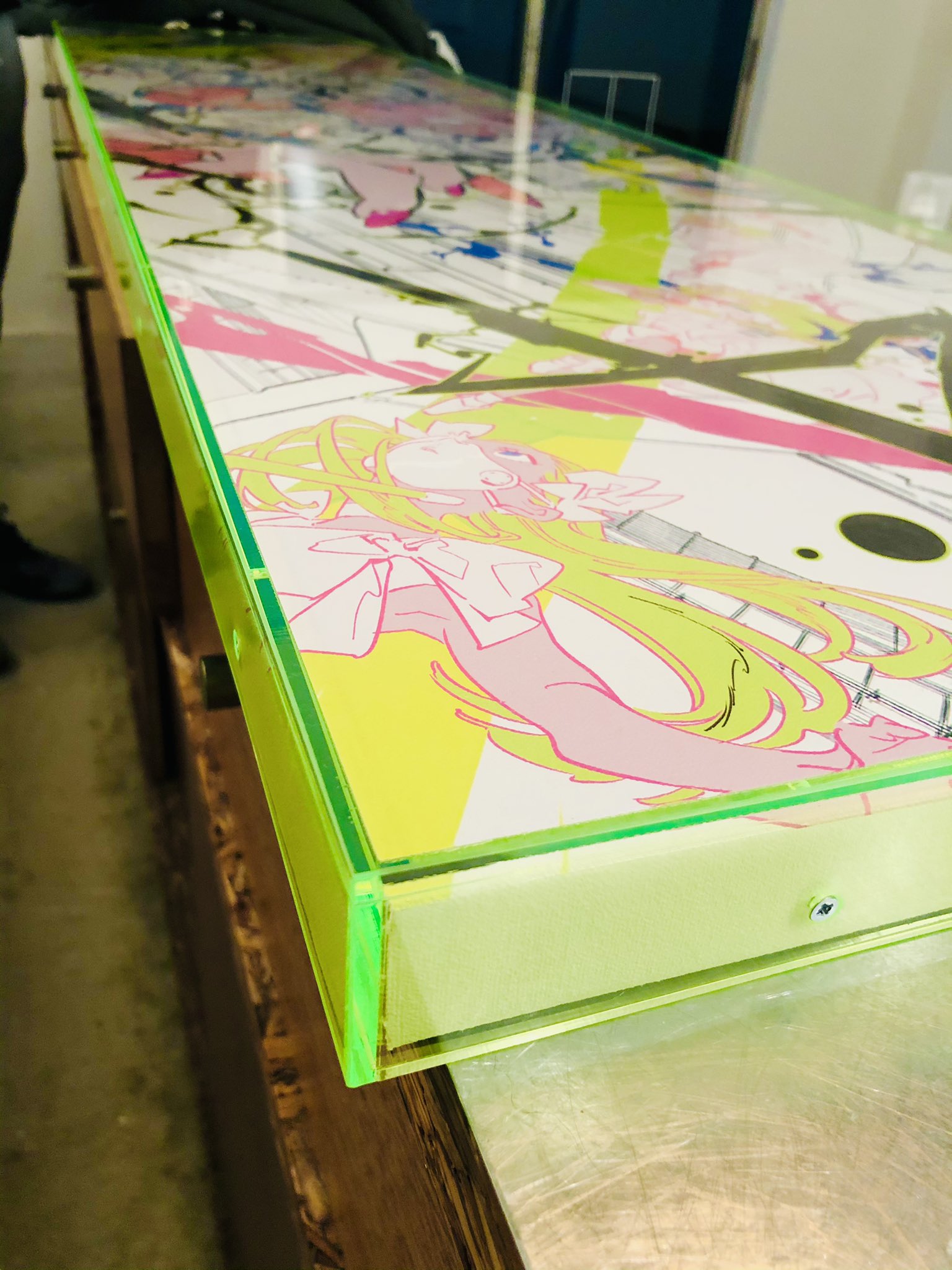 this is an image of a glass tabletop with an animestyle illustration. the illustration features a girl with long green hair wearing a pink and white outfit. the tabletop is framed by a thick, bright green border and is displayed on a wooden table. the image is brightly lit and taken from a high angle.