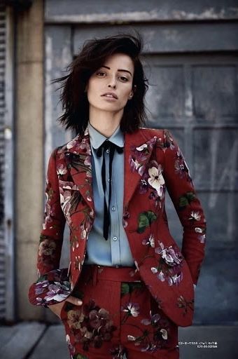 a medium shot of a woman in a red floral suit and light blue blouse with a black bow tie, posing in front of a grey wall. she is looking directly at the camera with a serious expression, and her dark hair is windswept. her left hand is in her pocket. the suit is covered in a floral pattern, and she is dressed in professional attire.