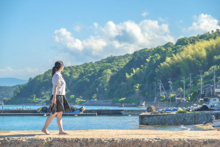 a young woman in a school uniform walks barefoot on a pier, holding her shoes in her hand. the background features a beautiful seascape with blue water, trees, and a clear sky with fluffy clouds. a small village and hills can be seen in the distance.
