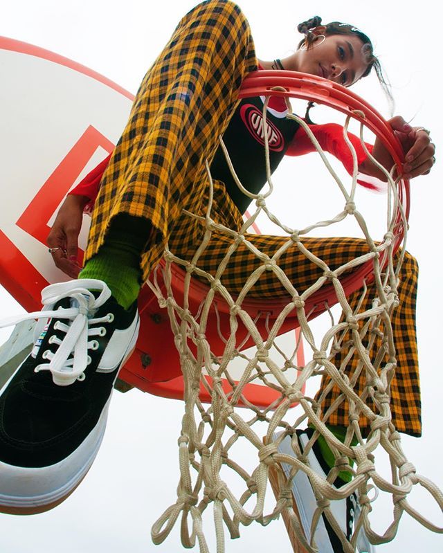 a young woman wearing black and white sneakers and yellow plaid pants hangs off of a basketball hoop with her legs crossed and her arms outstretched.  her hair is braided.