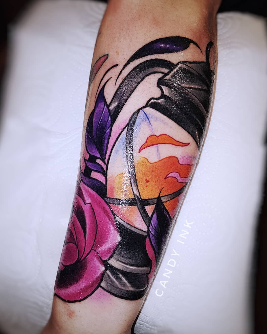 a forearm tattoo of a lantern with flames inside and a rose at the bottom. the tattoo has bold lines and vibrant colors, and is done in a neo traditional style. the tattoo artist's signature, "candy ink", is visible in black ink at the bottom right of the tattoo.