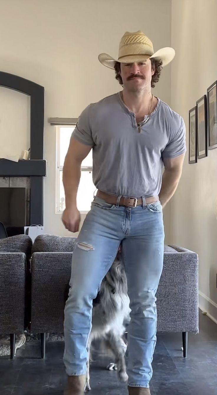 a muscular man wearing a cowboy hat and light blue shirt smiles while standing in a living room with his border collie. he is wearing light wash jeans and a brown belt. the man is casually dressed and relaxed, seemingly comfortable in his home environment. the dog is standing mostly behind him, partially hidden. the room's background features grey upholstered furniture and a fireplace.